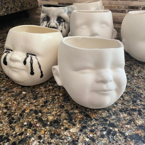 May include: A selection of ceramic baby-face planters. Some are plain white, others have black tear accents. The planters have a smooth, glossy finish and are designed with open tops for planting. They are suitable for indoor use.