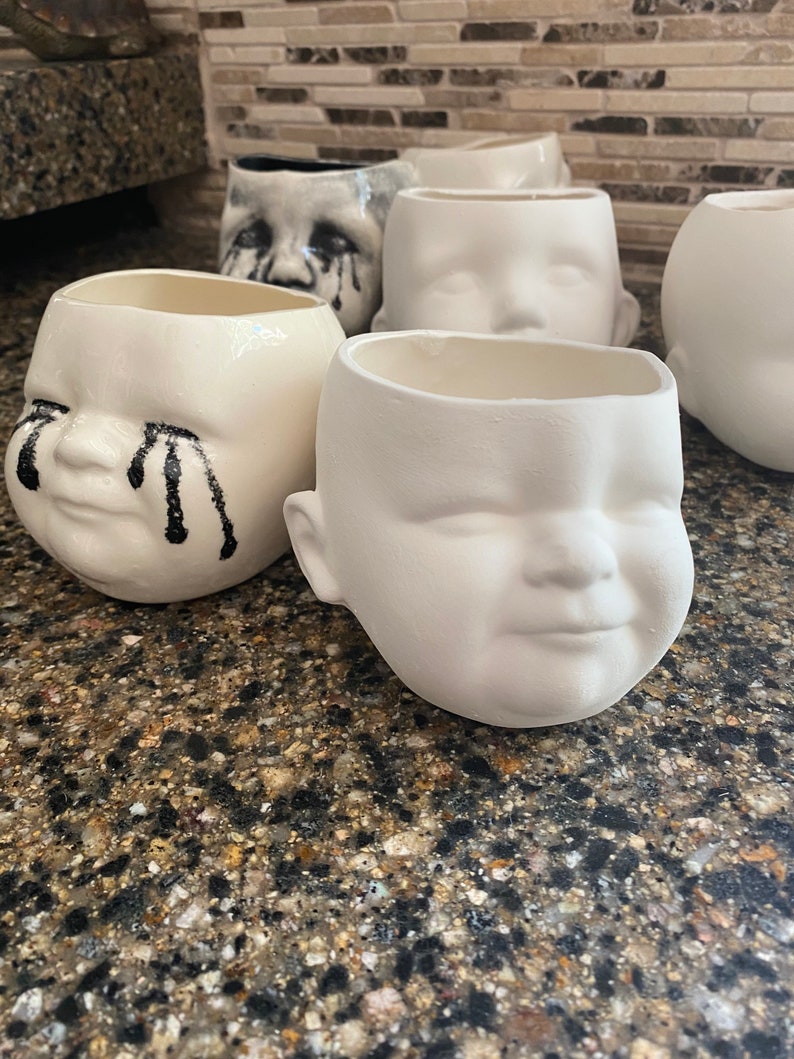 May include: A selection of ceramic baby-face planters. Some are plain white, others have black tear accents. The planters have a smooth, glossy finish and are designed with open tops for planting. They are suitable for indoor use.