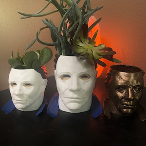 May include: Three white and brown ceramic planters shaped like the face of Michael Myers from the Halloween movies. The planters are filled with green succulents.