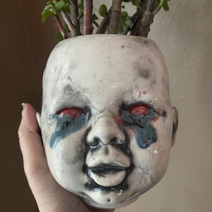 Puede incluir: Una maceta de cerámica blanca con forma de cabeza de bebé, con detalles rojos y azules alrededor de los ojos. La maceta está llena de plantas verdes. El diseño tiene una cualidad inquietante, pero artística.