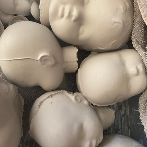May include: A group of unpainted, off-white ceramic doll heads. The heads are in different orientations, some with visible ears and closed eyes. The unfinished state indicates they are ready for painting or further customisation. The heads are in a container.