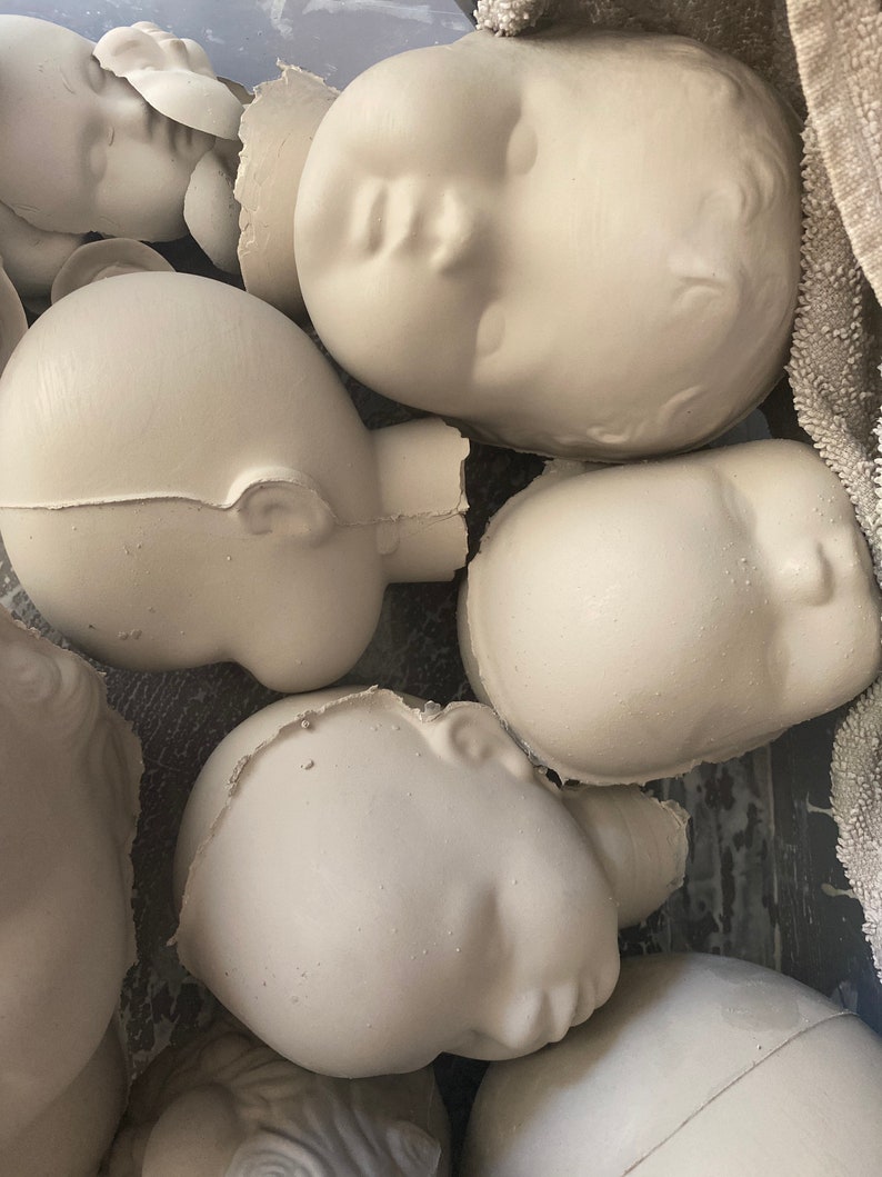 May include: A group of unpainted, off-white ceramic doll heads. The heads are in different orientations, some with visible ears and closed eyes. The unfinished state indicates they are ready for painting or further customisation. The heads are in a container.
