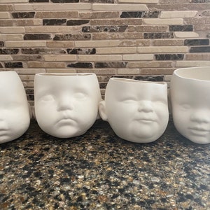 Puede incluir: Cuatro macetas de cerámica blanca con forma de caras de bebé. Cada maceta tiene una expresión facial diferente, incluyendo una cara neutra, triste y sonriente. Las macetas están dispuestas en fila sobre una superficie oscura y moteada.
