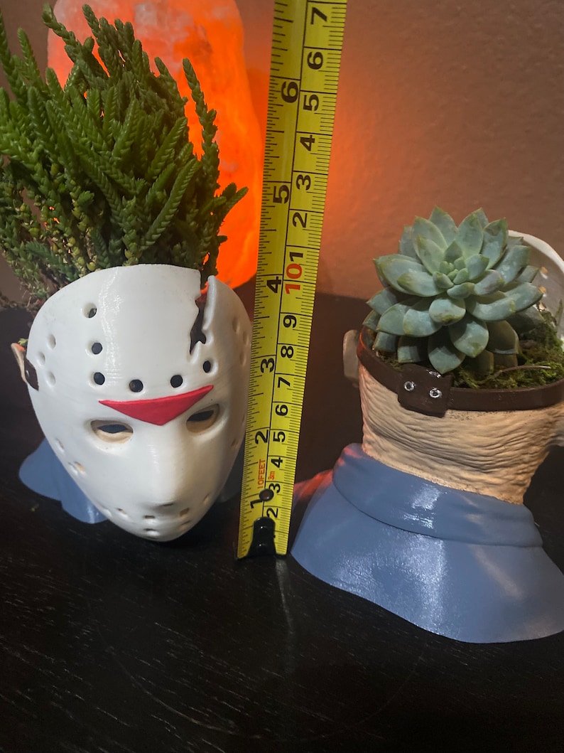 Puede incluir: Dos macetas de cer&aacute;mica blanca con forma de m&aacute;scara de hockey y cabeza humana, con suculentas verdes creciendo en su interior. Las macetas miden aproximadamente 15 cm de alto.