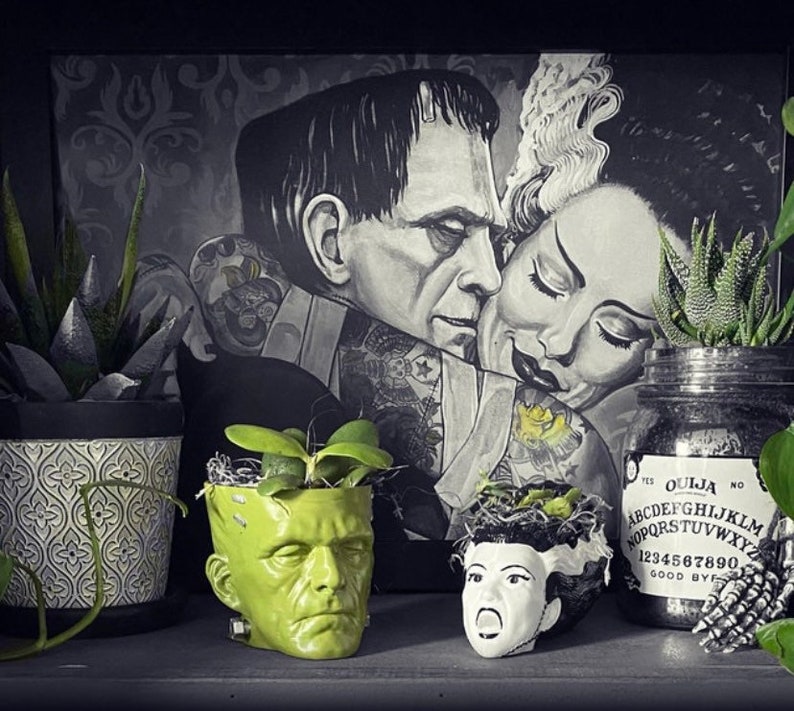 May include: A black and white illustration of Frankenstein's monster and his bride. The illustration is framed and hanging on a wall. There are three potted plants in front of the illustration, one with a green ceramic pot shaped like a monster's head, one with a white ceramic pot with a floral pattern, and one in a clear glass jar with a black and white Ouija board label.