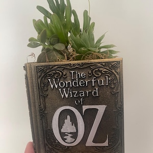 May include: A decorative planter shaped like a book, featuring the text "The Wonderful Wizard of OZ" in white lettering. The planter is filled with green succulents and has a bronze-colored finish.