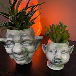 May include: Two ceramic face planters, one large and one small, each holding a succulent plant. The planters are painted in shades of grey and green, with detailed facial features. The larger planter has a smiling expression.