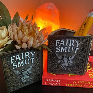 May include: Two decorative book-shaped pen holders with the text "FAIRY SMUT" and a fairy illustration. One holds pencils, the other flowers. The holders are on a table with a book and a glowing orange light in the background.