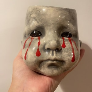 Pode incluir: Um vaso de cerâmica em forma de rosto de criança com lágrimas pintadas de vermelho. O rosto tem uma expressão triste e é pintado em tons de cinza.