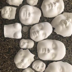 Op de afbeelding: Een verzameling van 14 witte gipsen poppenkopvormen. De vormen zijn van verschillende groottes en vormen, met verschillende gezichtsuitdrukkingen.