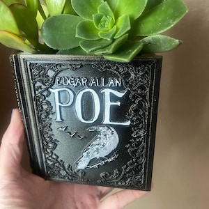 Może przedstawiać: Czarny, książkowy pojemnik na rośliny z napisem "Edgar Allan Poe" i ilustracją białego kruka. Pojemnik jest wypełniony zielonymi sukulentami.