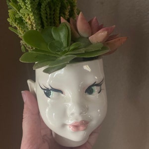 May include: A white ceramic planter shaped like a human head with painted facial features including black winged eyeliner, green eyes, and pink lips. The planter is filled with a variety of green and pink succulent plants growing from the top.
