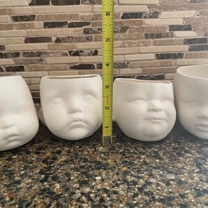 May include: Four white ceramic mugs shaped like baby faces. The mugs are arranged in a row, with a measuring tape next to them. The mugs are approximately 7.6 cm tall.