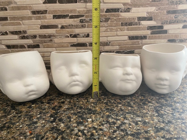 May include: Four white ceramic mugs shaped like baby faces. The mugs are arranged in a row, with a measuring tape next to them. The mugs are approximately 7.6 cm tall.