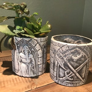 Puede incluir: Dos macetas de cerámica con temática medieval. Una maceta presenta una figura blanca de un hombre con barba y bastón, mientras que la otra maceta presenta una espada y un hacha blancas. Ambas macetas tienen una textura de piedra gris.