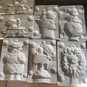 May include: A selection of unpainted plaster moulds depicting festive Christmas characters. Designs include a bear on a sledge, a star with a face, and a bear holding a sign that says "North Pole".