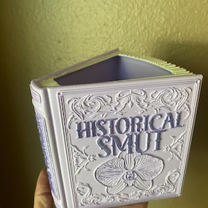 May include: A lavender-colored book-shaped container with the words "HISTORICAL SMUT" and an orchid design. The container has an open top and is held by a hand.