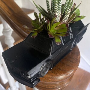 May include: A black car-shaped planter with a variety of succulents growing inside. The planter is made of a smooth, matte black plastic and has a realistic car design.