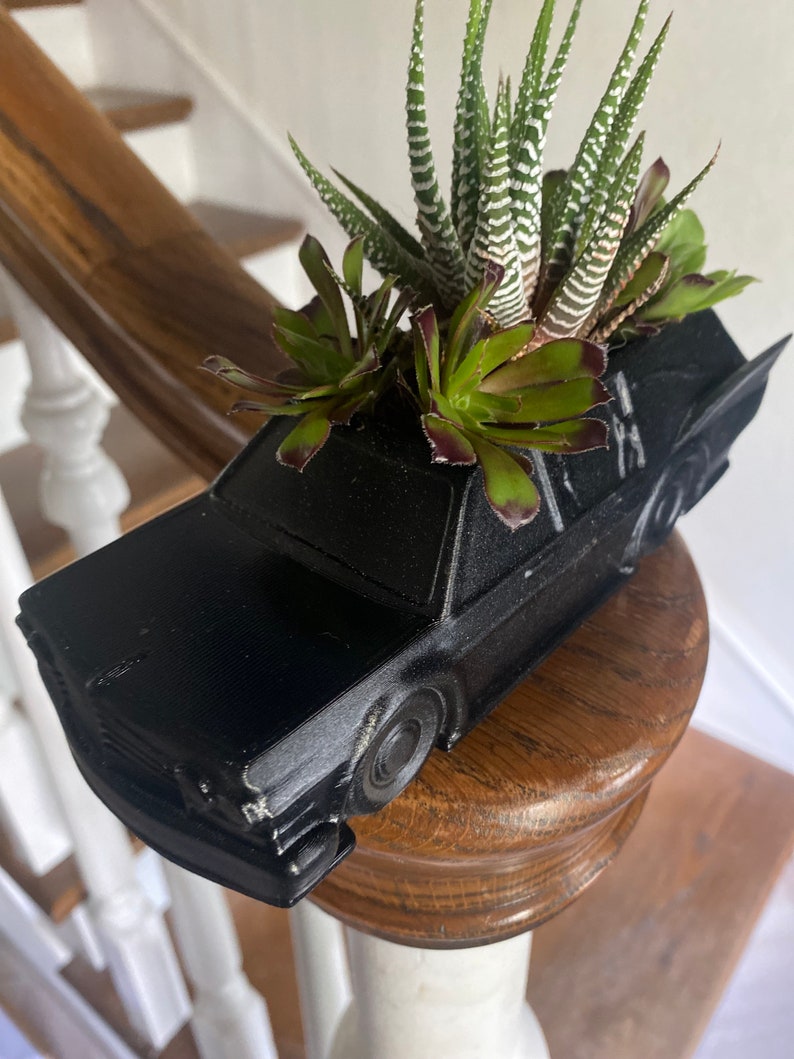 May include: A black car-shaped planter with a variety of succulents growing inside. The planter is made of a smooth, matte black plastic and has a realistic car design.