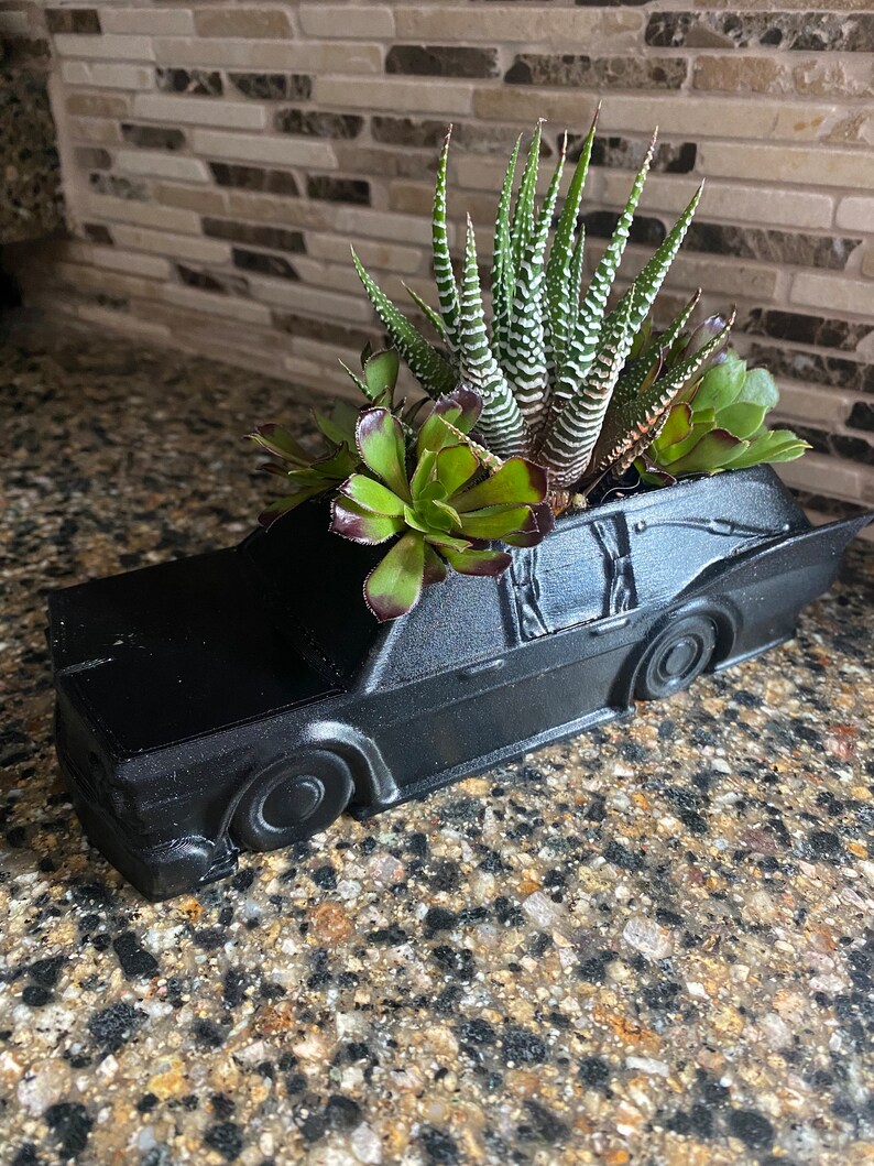 May include: A black car-shaped planter with a variety of succulents growing inside. The planter is made of a smooth, matte black material and has a realistic car design.