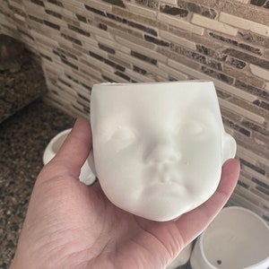 May include: A white ceramic planter in the shape of a baby's face, with detailed facial features. The planter has a hollow interior for plants or decorative items. Other planters are visible in the background.