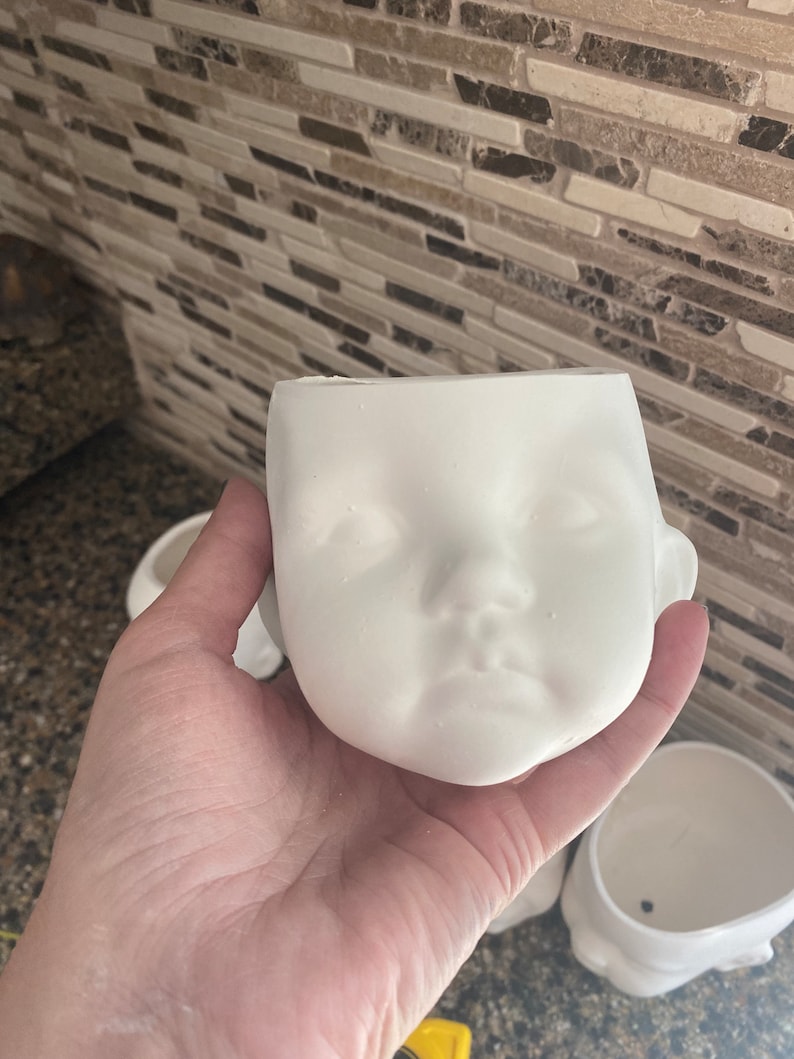 May include: A white ceramic planter in the shape of a baby's face, with detailed facial features. The planter has a hollow interior for plants or decorative items. Other planters are visible in the background.