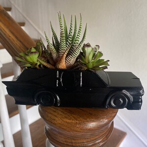 May include: A black car-shaped planter with a variety of succulents growing inside. The planter is sitting on a wooden railing.