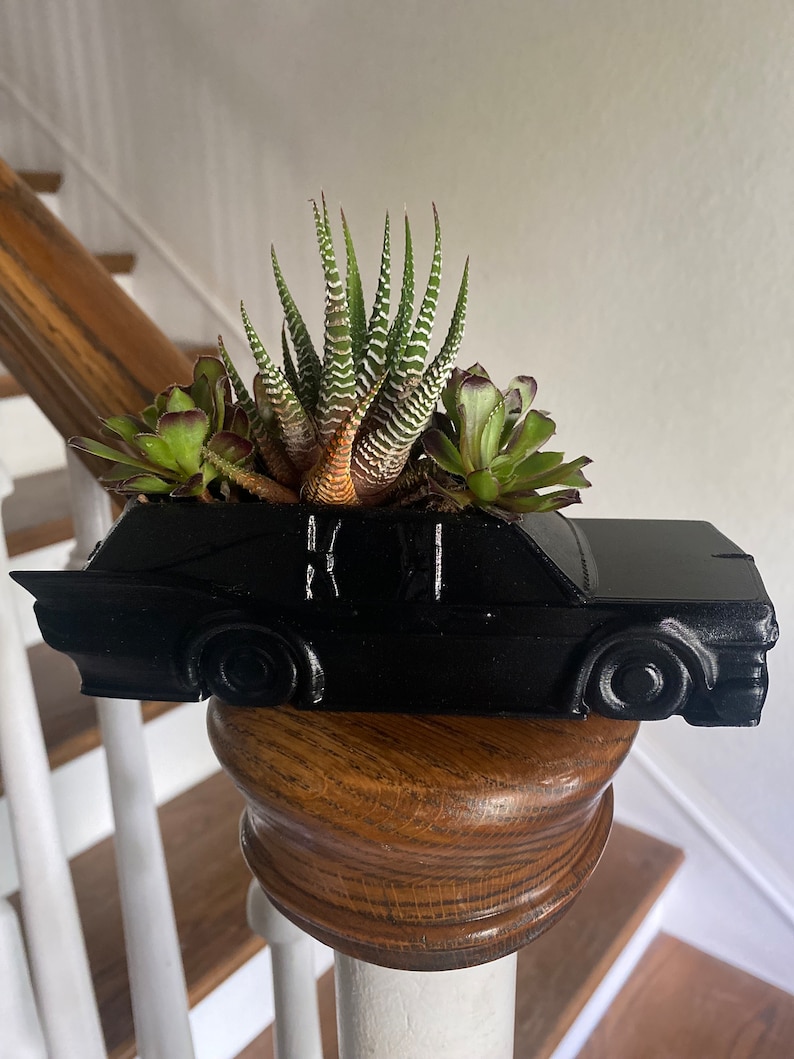 May include: A black car-shaped planter with a variety of succulents growing inside. The planter is sitting on a wooden railing.