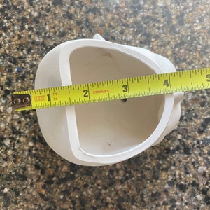 May include: A white ceramic skull-shaped planter with a 10 cm opening, measured with a yellow tape measure. The planter has a smooth surface and a small drainage hole. The skull has a detailed design.