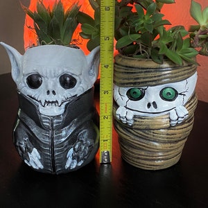 K&ouml;nnte beinhalten: Zwei Halloween-Keramik-Pflanzgef&auml;&szlig;e, eines im Vampir-Design und das andere als Mumie. Der Vampir-Pflanztopf ist etwa 15 cm hoch, mit einem schwarzen Kragen. Der Mumien-Pflanztopf ist etwa 13 cm hoch. Beide enthalten gr&uuml;ne Sukkulenten.