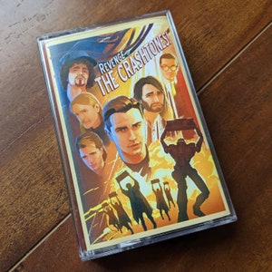Könnte beinhalten: Eine Kassette mit einer gelben und roten Illustration einer Band namens "The Crashitones". Die Bandmitglieder sind in einem Retro-Stil mit einem Roboter im Vordergrund dargestellt. Der Text "Revenge of The Crashitones!" ist auf der Kassette aufgedruckt.