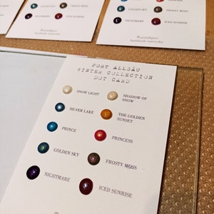 Könnte beinhalten: Eine weiße Karte mit dem Text "Port Allgäu Winter Collection Dot Card" und einer Farbkarte mit 10 Aquarellpunkten. Die Punkte sind mit Namen wie "Schneelicht", "Schatten des Schnees", "Silbersee", "Der goldene Sonnenuntergang", "Prinz", "Prinzessin", "Goldener Himmel", "Frostiger Moos", "Alptraum" und "Eisiger Sonnenaufgang" beschriftet.