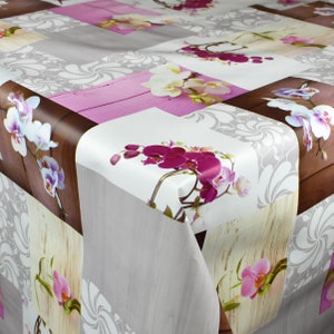 Op de afbeelding: Een wit tafelkleed met een patchwork-ontwerp met roze, bruine en beige houtnerfpatronen. Het tafelkleed is versierd met verschillende orchideeën in tinten wit, roze en paars.