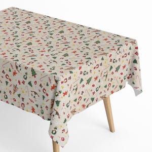 Op de afbeelding: Een rechthoekig tafelkleed met een feestelijk kerstpatroon. De gebroken witte stof is bedekt met herhaalde illustraties van kerstbomen, zuurstokken, rendieren en notenkrakers in rood, groen en geel.