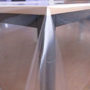 Könnte beinhalten: Eine transparente Plastiktischdecke, die über einen Holztisch gelegt wird. Die Tischdecke ist glatt und faltenfrei und hängt über die Seiten des Tisches.