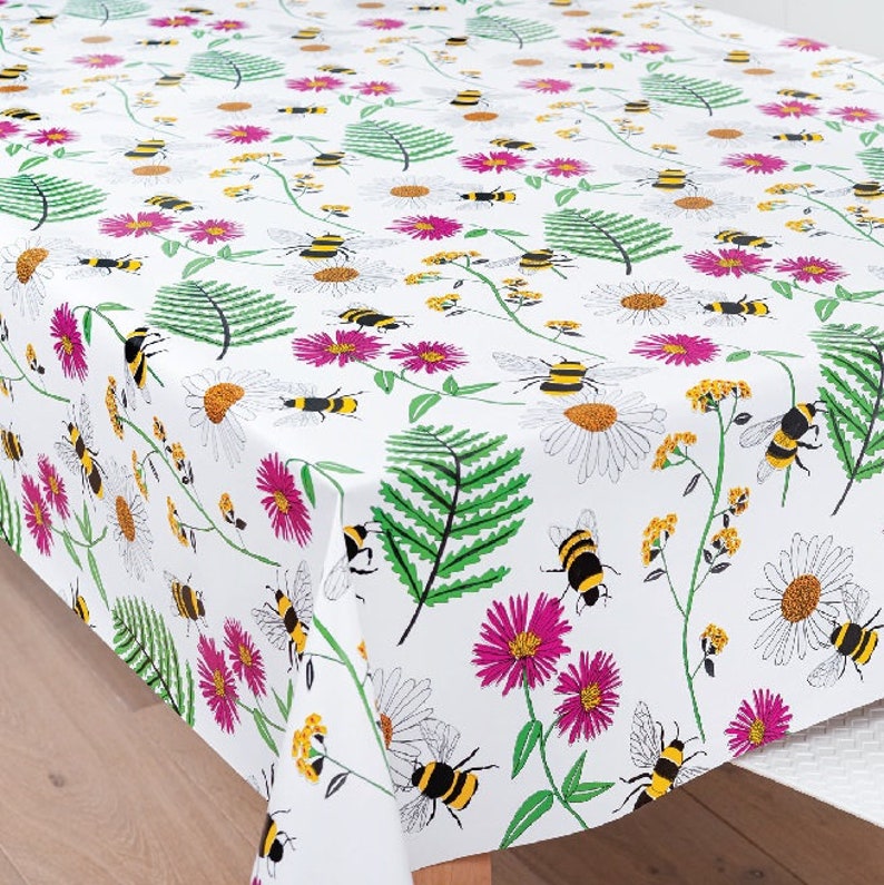 May include: White tablecloth with a repeating pattern of bees, flowers, and ferns. The bees are yellow and black, the flowers are pink and white, and the ferns are green.