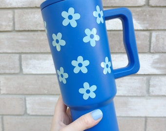 May include: A royal blue insulated tumbler with a handle and a matching straw, featuring a repeating pattern of light blue five-petaled flowers. The base of the tumbler displays the letters slm in a small, simple font.