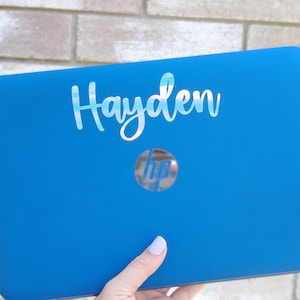 May include: A blue HP laptop with the name "Hayden" in a cursive font. The laptop has the HP logo in the center. The laptop is held by a person with light-colored nail polish.