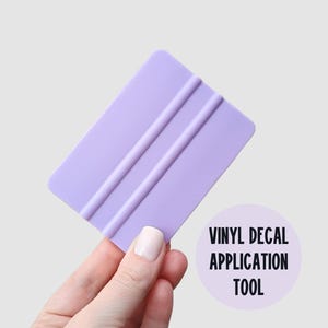 May include: A light purple vinyl decal application tool with three parallel ridges. The tool is held by a hand with a light-colored nail polish. Text on the image reads "VINYL DECAL APPLICATION TOOL".