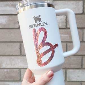 May include: White Stanley travel mug with a pink glitter letter "B" on the front. The mug has a black logo with a bear and the word "STANLEY" on it.