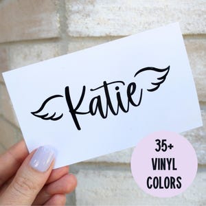 May include: A white rectangular card with the name "Katie" in black script, adorned with black wing designs. A pink circle below reads "35+ VINYL COLORS". The card is held against a brick wall.