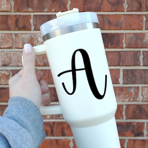 May include: White travel mug with a black letter A on the side. The mug has a handle and a lid.