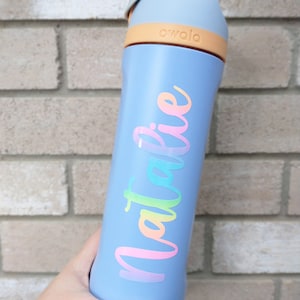 May include: A light blue Owala water bottle with a rainbow-colored "Natalie" decal. The bottle has a light orange band and a gray handle. The bottle is held in a hand against a brick wall.
