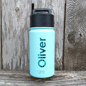 May include: A light blue water bottle with a black lid and handle. The name "Oliver" is printed vertically in teal on the bottle. The brand logo "slm" is printed below the name. The bottle is cylindrical and appears to be made of metal.