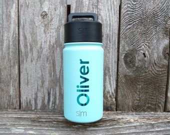 May include: A light blue water bottle with a black lid and handle. The name "Oliver" is printed vertically in teal on the bottle. The brand logo "slm" is printed below the name. The bottle is cylindrical and appears to be made of metal.