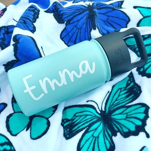 May include: A light blue water bottle with a black handle and the name "Emma" in white script. The bottle is resting on a white towel with blue and teal butterfly patterns. The bottle has a matte finish.