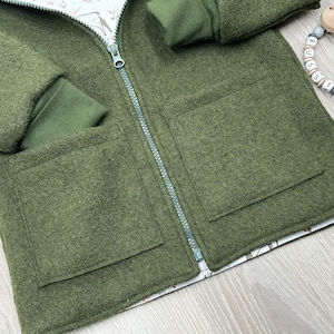 Peut inclure: Gros plan sur une veste en laine verte avec une fermeture éclair complète et deux poches avant. La veste a des poignets côtelés verts et une doublure à motifs. Un anneau de dentition en bois avec des perles lettrées est dans le coin supérieur droit.