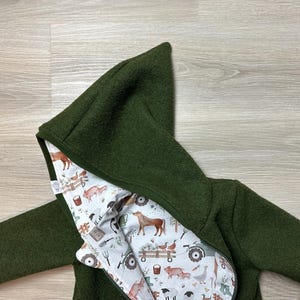 Könnte beinhalten: Dunkelgrüne Kapuzenjacke mit durchgehendem Reißverschluss. Das Innenfutter zeigt einen Bauernhoftierdruck in Braun, Weiß und Grau. Die Jacke ist aus einem weichen, strukturierten Material gefertigt, und die Kapuze ist vollständig gefüttert.