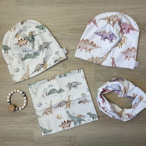 Könnte beinhalten: Ein Set Baby-Accessoires mit Dinosaurier-Motiv. Das Set enthält zwei weiße Mützen, einen Halstuch und einen passenden Loop-Schal. Die Artikel sind mit farbenfrohen Dinosaurier-Illustrationen in Grün-, Braun- und Rosatönen verziert.