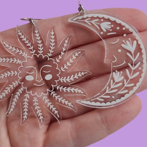 May include: A pair of clear acrylic earrings with white designs. One earring is a sun with a face and the other is a crescent moon with floral details.